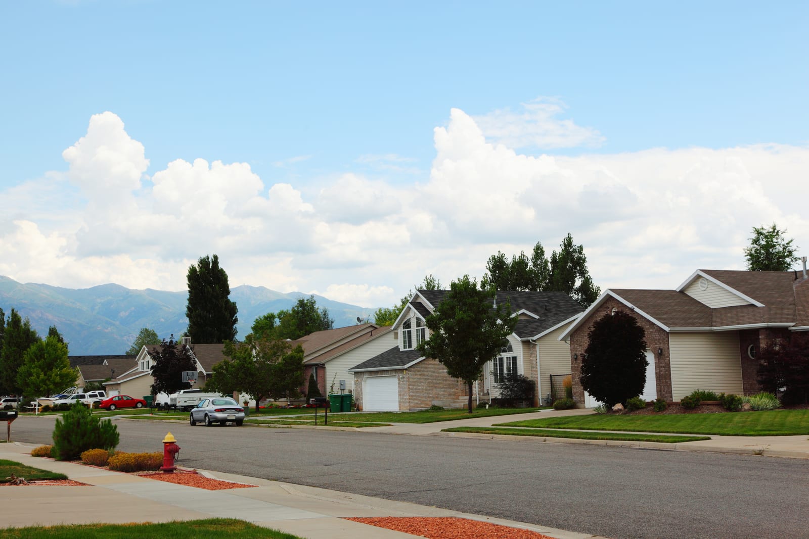 A suburban residential street showing middle-class homes with front yards and sidewalks on a clear day