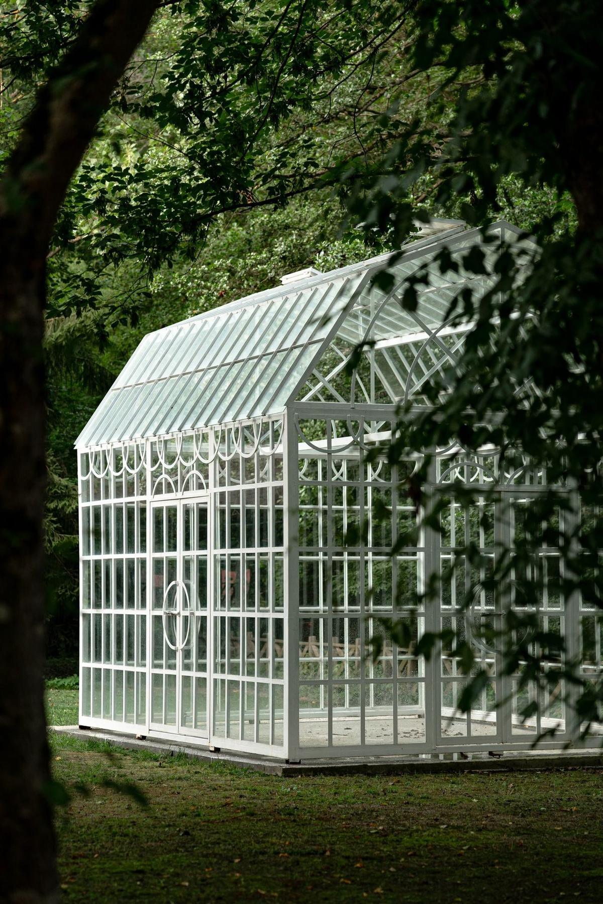 A Victorian-style glass greenhouse surrounded by lush greenery in a garden park, dark aluminum framing visible through the trees